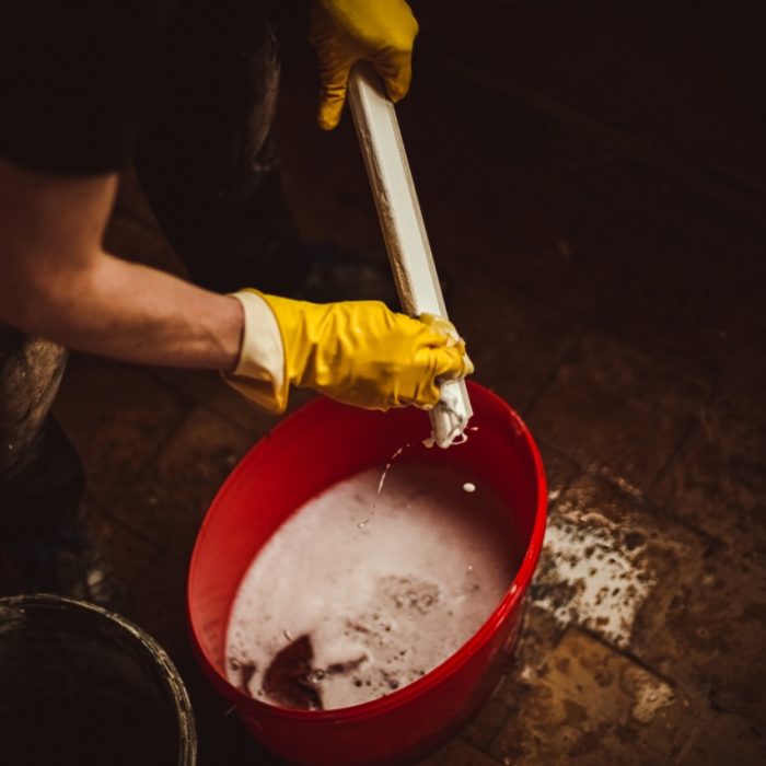 One young Caucasian man in yellow gloves washes with water and soapy water using a sponge a narrow part of a plastic window frame in a red bucket in a house where renovation is underway, close-up side view with selective focus. Concept of home renovation and washing window frames.