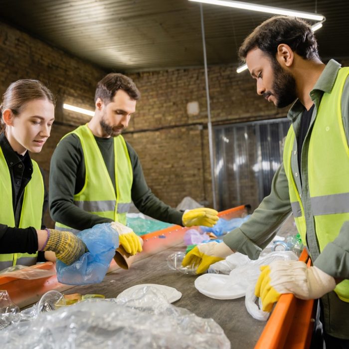 Multiethnic workers in protective vests and gloves taking trash from conveyor while working together in waste disposal station at background, garbage sorting and recycling concept