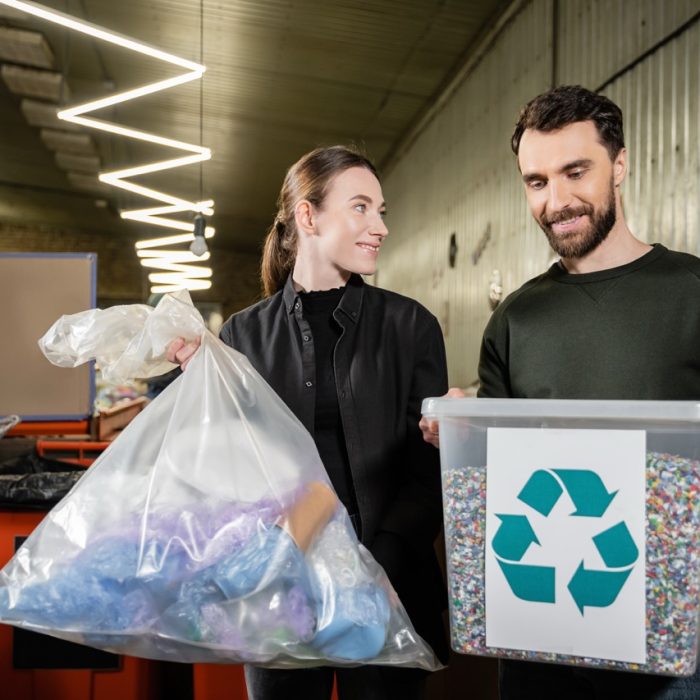 Smiling volunteer holding trash bag near man with bin and recycle sign in blurred waste disposal station at background, garbage sorting and recycling concept